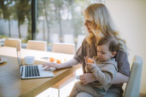 A mother works on a laptop at home while holding her baby, showcasing remote working and parenting.